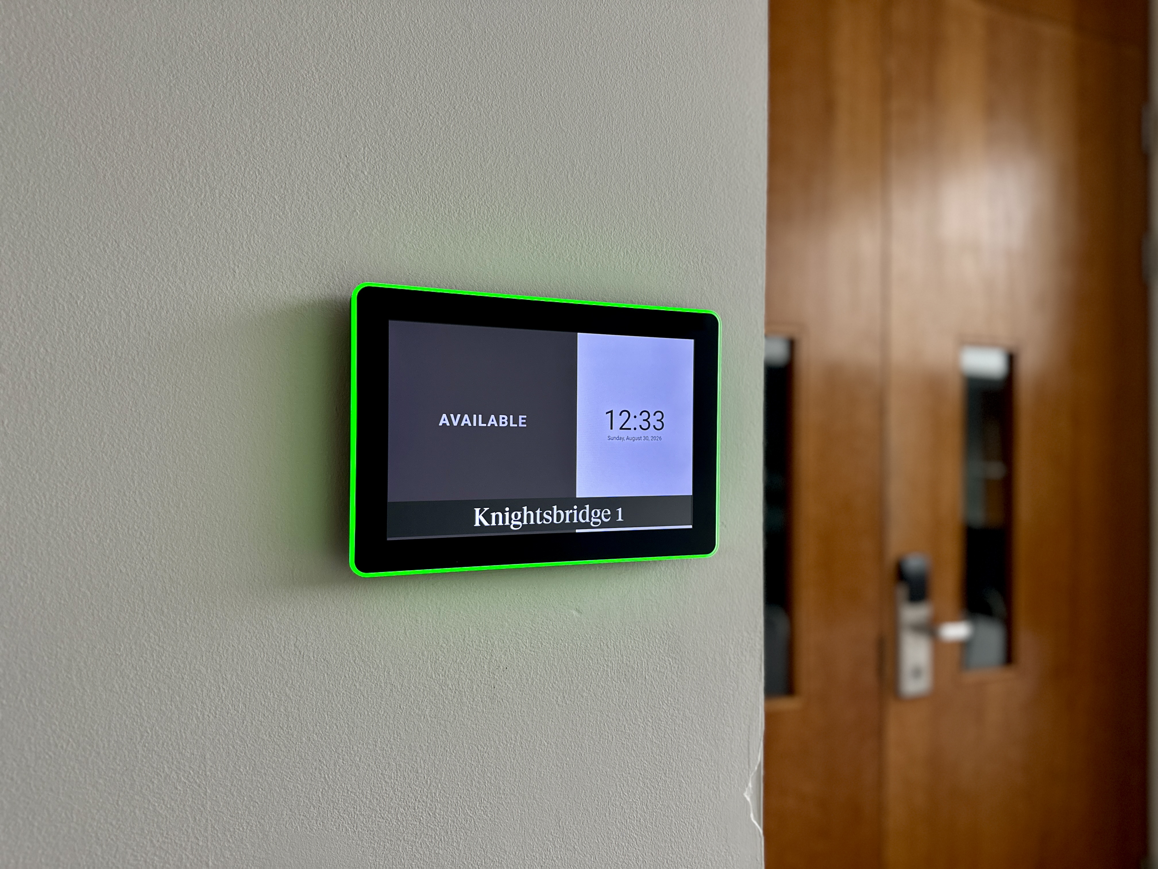 Wall-mounted tablet with a bright green bezel showing 'AVAILABLE' and 'Knightsbridge 1' beside a wooden door panelled wall.