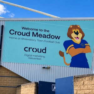Teal banner on a stadium wall reads 'Welcome to the Croud Meadow, Home of Shrewsbury Town Football Club' with a cartoon lion mascot against a blue sky.