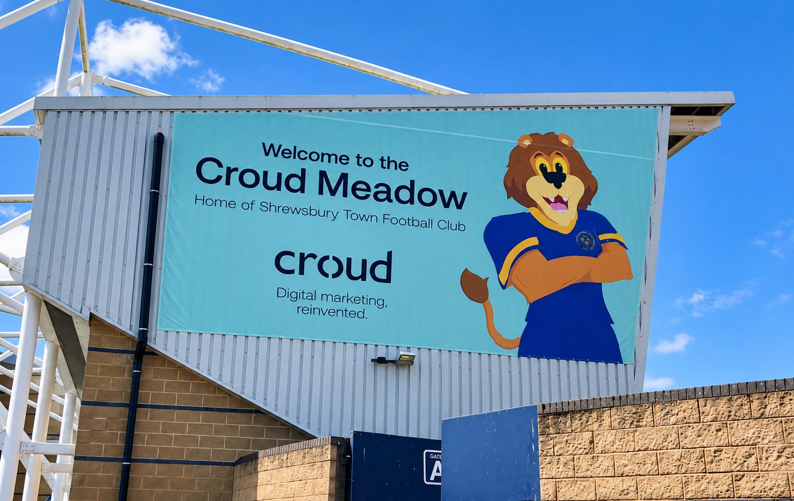 Teal banner on a stadium wall reads 'Welcome to the Croud Meadow, Home of Shrewsbury Town Football Club' with a cartoon lion mascot against a blue sky.