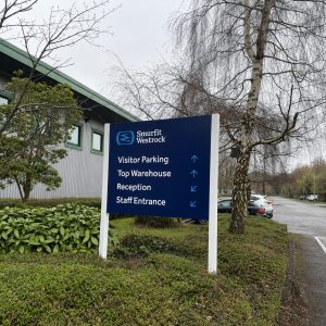Blue directional sign at Smurfit Westrock listing Visitor Parking, Top Warehouse, Reception, and Staff Entrance with arrows, in a landscaped lot.