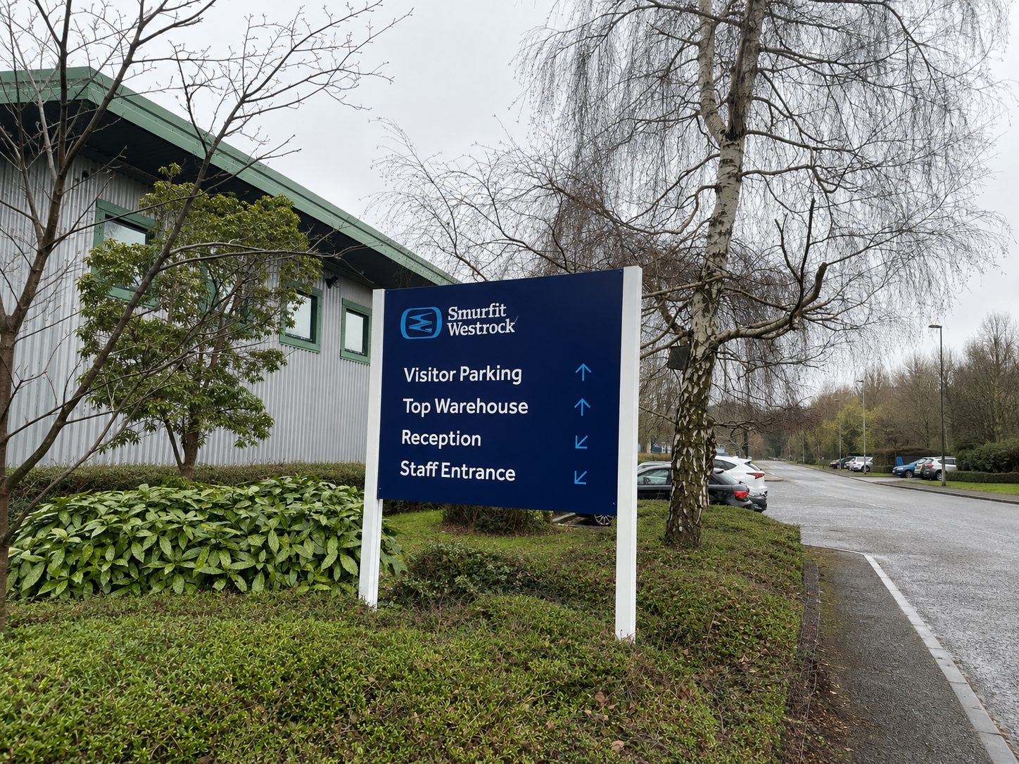 Blue directional sign at Smurfit Westrock listing Visitor Parking, Top Warehouse, Reception, and Staff Entrance with arrows, in a landscaped lot.