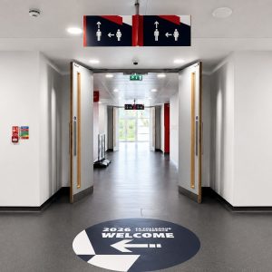 Long, bright hallway with double doors open to a sunlit space; overhead signs and a circular floor graphic reading '2026 WELCOME' with an arrow.