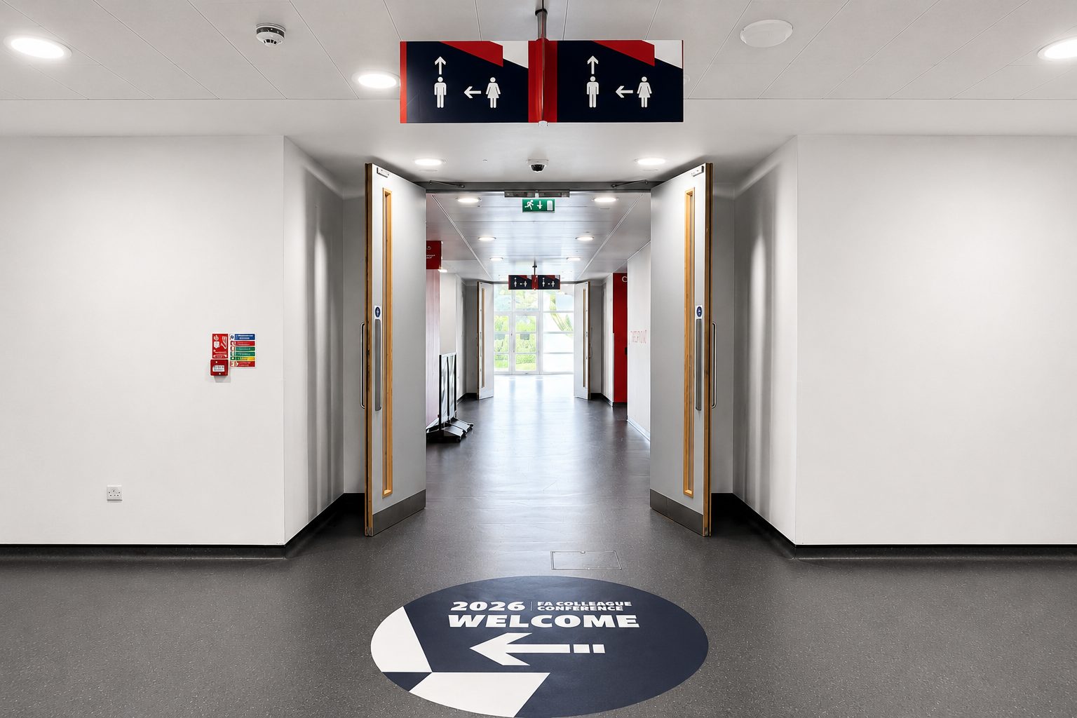 Long, bright hallway with double doors open to a sunlit space; overhead signs and a circular floor graphic reading '2026 WELCOME' with an arrow.