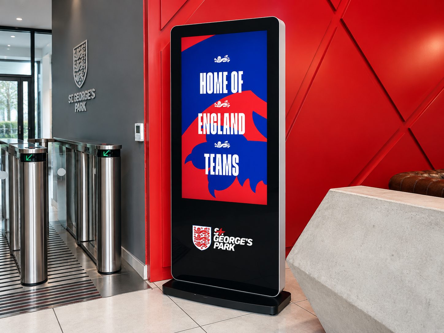 Tall digital sign displaying 'HOME OF ENGLAND TEAMS' with England crest, in a hallway near turnstiles at St George's Park