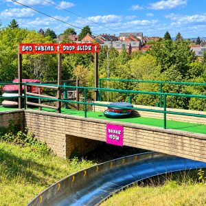 Outdoor tubing slide setup with stacked colorful inner tubes, green railings, and a bright 'Snow Tubing Drop Slide' sign against a suburban backdrop.