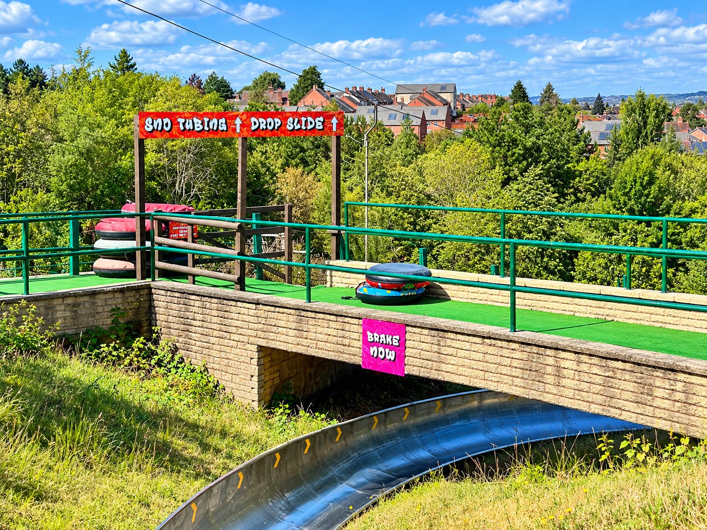Outdoor tubing slide setup with stacked colorful inner tubes, green railings, and a bright 'Snow Tubing Drop Slide' sign against a suburban backdrop.