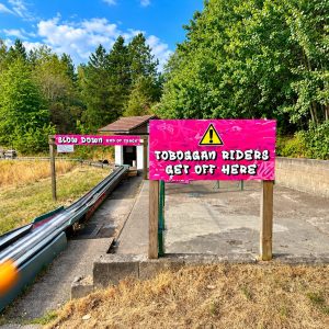 Bright pink sign says 'TOBOGAN RIDERS GET OFF HERE' at the end of a small toboggan track in a wooded outdoor area.