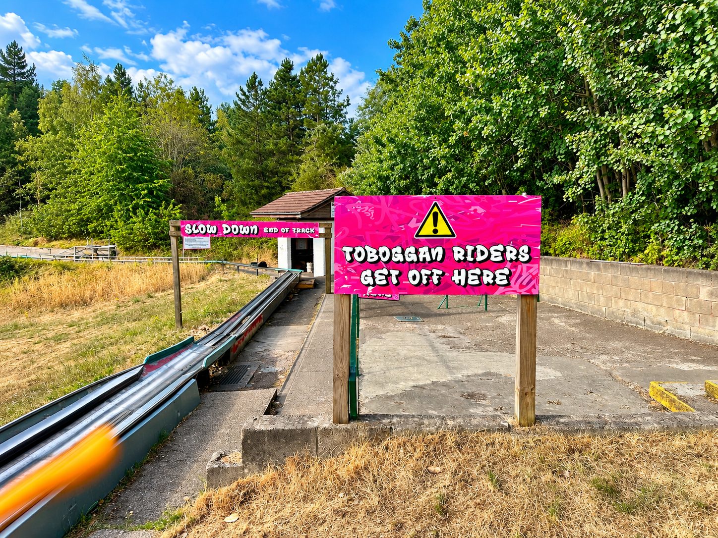 Bright pink sign reading 'TOBOGGAN RIDERS GET OFF HERE' at the end of a small track, with a yellow warning triangle above, outdoors among trees and a hut in the background.