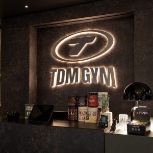Reception counter in a dark marble lobby with a glowing TDM GYM logo on the wall behind and fitness products displayed on the counter.