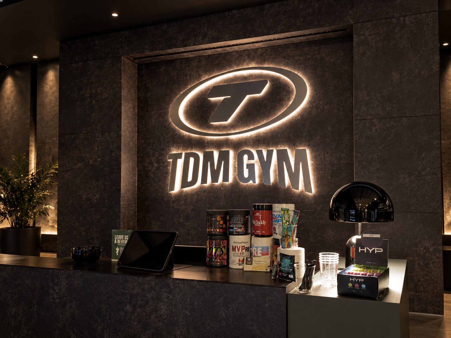 Reception counter in a dark marble lobby with a glowing TDM GYM logo on the wall behind and fitness products displayed on the counter.