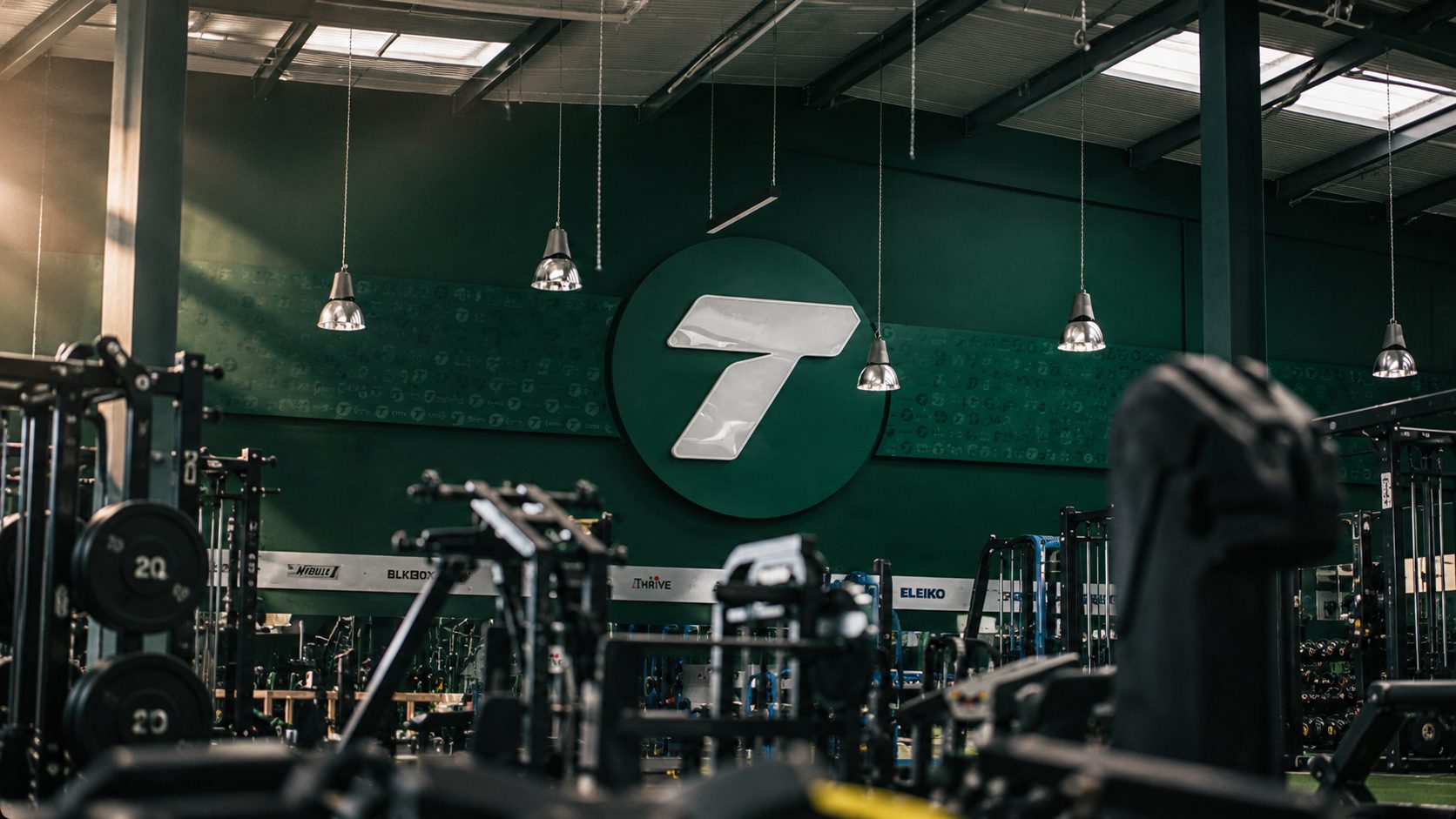 Gym interior with heavy weights and racks; large green wall logo featuring a white circular emblem with a bold '7' in the center.