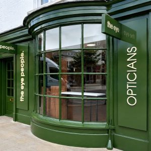 Green storefront of an eyewear shop with a large curved bay window, vertical white lettering, and signs reading 'the eye' and 'people' on the green facade.