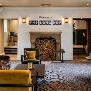 Cozy pub lounge with a stone wall, fireplace with stacked wood, and a sign reading 'Welcome to THE LONG HOP' above the mantel. Warm seating around a round table and patterned carpet.