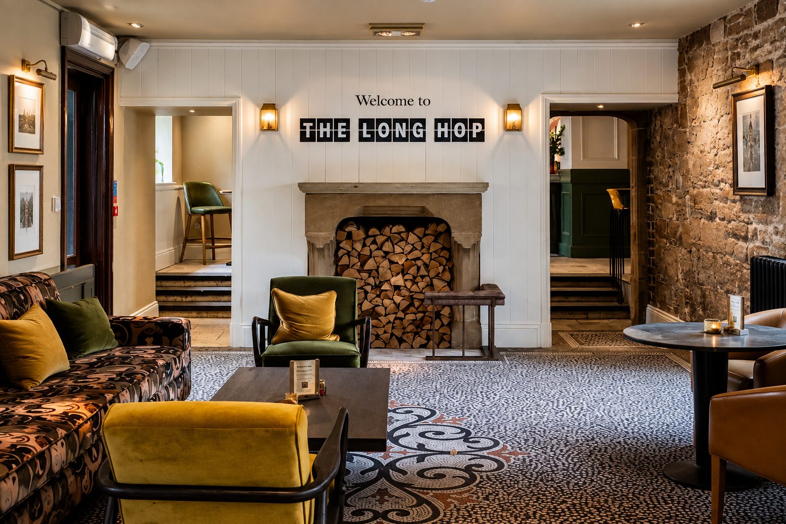 Cozy pub lounge with a stone wall, fireplace with stacked wood, and a sign reading 'Welcome to THE LONG HOP' above the mantel. Warm seating around a round table and patterned carpet.