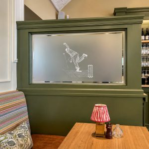 Green paneled cafe/bar area with a frosted glass panel featuring a bowler illustration; striped bench seating and a wooden table with a red-striped lamp nearby.
