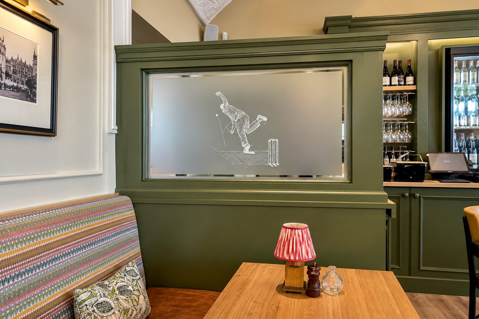 Green paneled cafe/bar area with a frosted glass panel featuring a bowler illustration; striped bench seating and a wooden table with a red-striped lamp nearby.