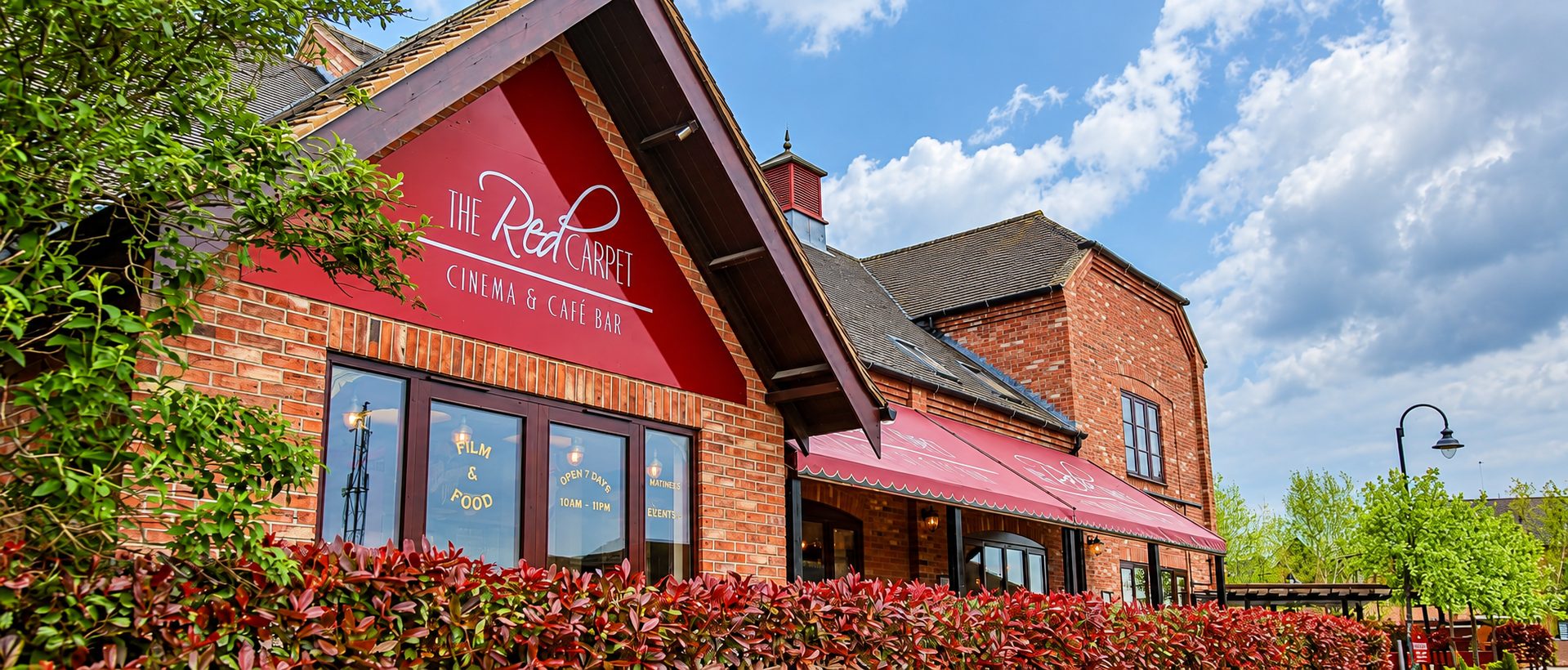 Exterior of a red-brick cinema and café bar named 'The Red Carpet' with a red awning and lush greenery in the foreground.