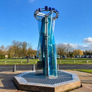 Tall glass sculpture in a circular plaza, sunlit park with blue sky behind it.