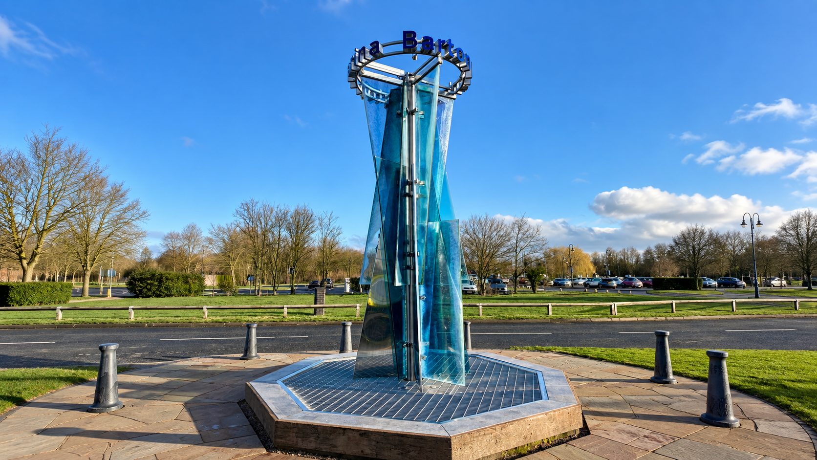 Tall glass sculpture in a circular plaza, sunlit park with blue sky behind it.