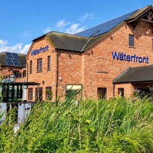 Brick waterfront building with blue 'Waterfront' sign, solar panels on the roof, and a boat moored alongside on a sunny day.