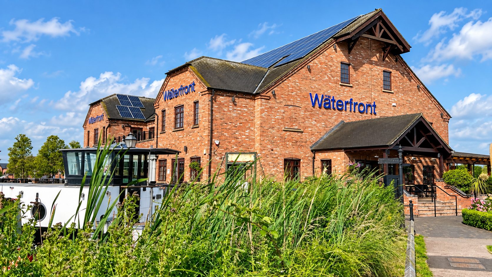 Brick waterfront building with blue 'Waterfront' sign, solar panels on the roof, and a boat moored alongside on a sunny day.
