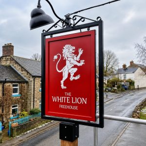 Red pub sign reading 'The White Lion Freehouse' with a white lion illustration, mounted over a street in a small village.