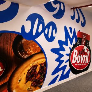 Bright wall mural advertising Bovril beef paste, featuring a large bottle cutout and close-up meal photos on circular frames.