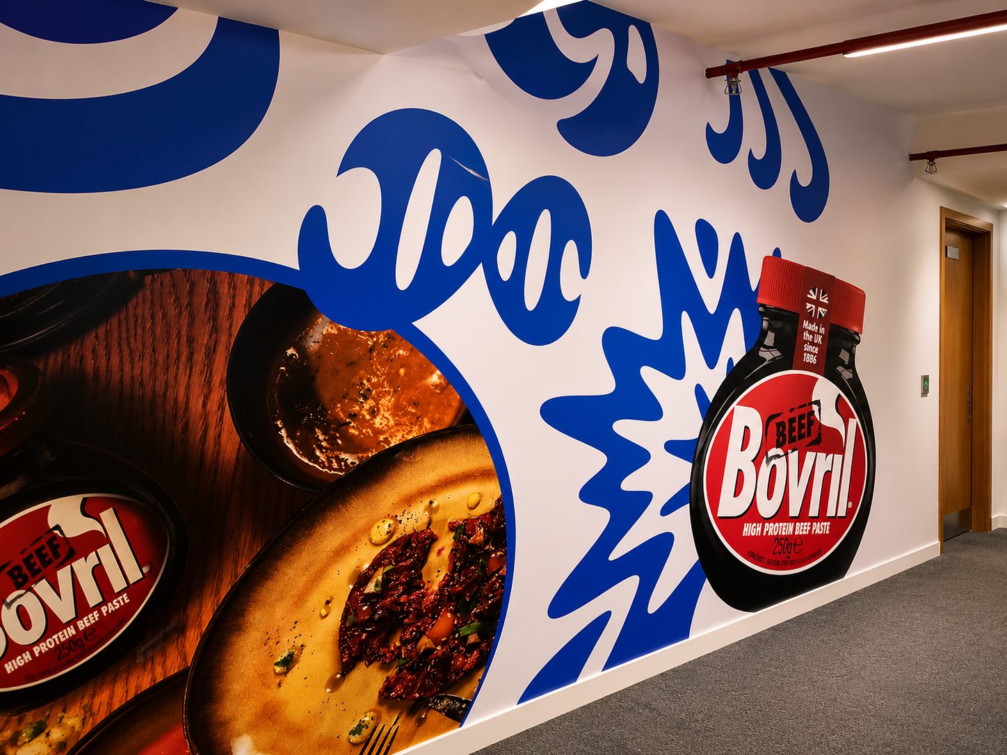 Bright wall mural advertising Bovril beef paste, featuring a large bottle cutout and close-up meal photos on circular frames.