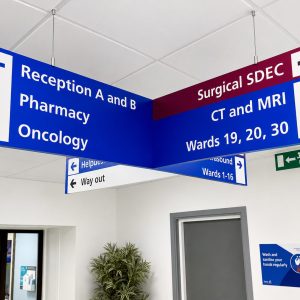 Large blue-and-white hanging hospital sign directing to Reception A and B, Pharmacy, Oncology, and imaging areas like CT/MRI and wards 19–30.