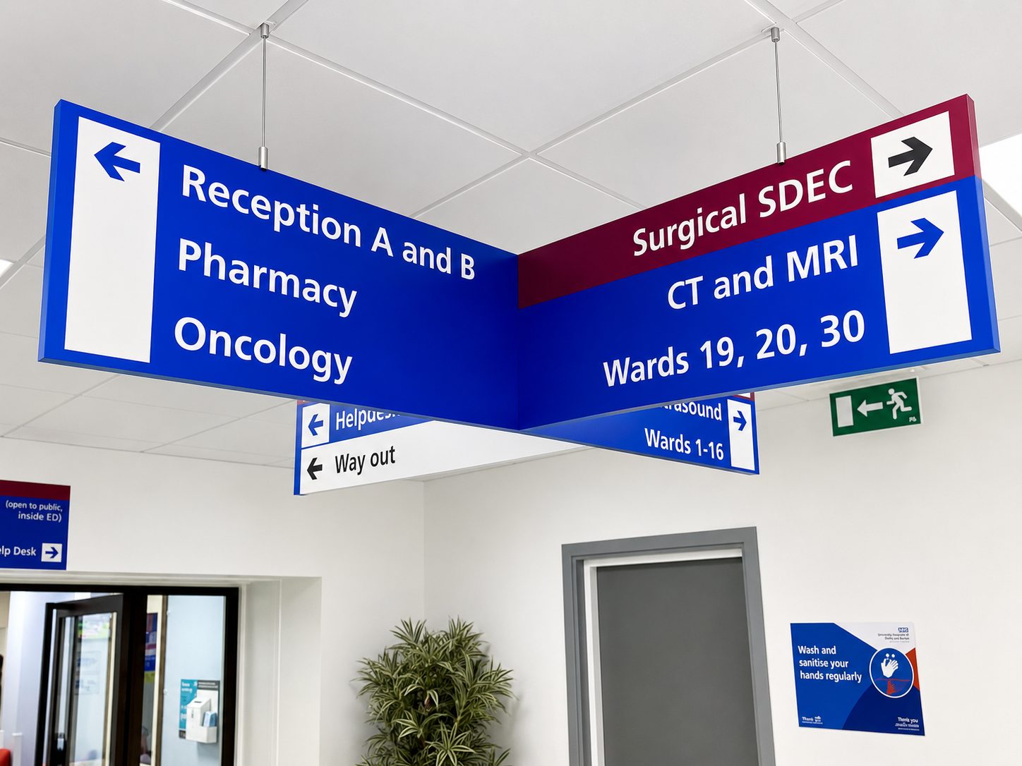 Large blue-and-white hanging hospital sign directing to Reception A and B, Pharmacy, Oncology, and imaging areas like CT/MRI and wards 19–30.