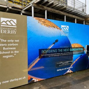 Construction-site barrier with University of Derby banners, blue mosaic artwork, and the slogan 'Powering the next generation' under construction scaffolding.
