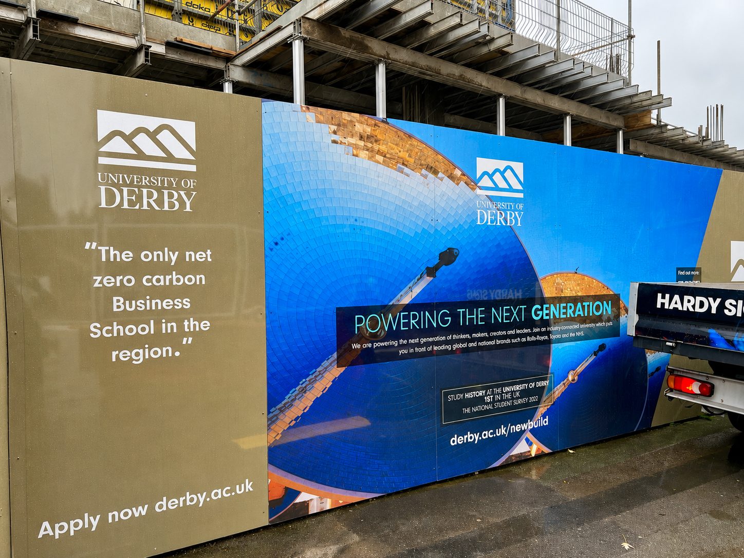 Construction-site barrier with University of Derby banners, blue mosaic artwork, and the slogan 'Powering the next generation' under construction scaffolding.
