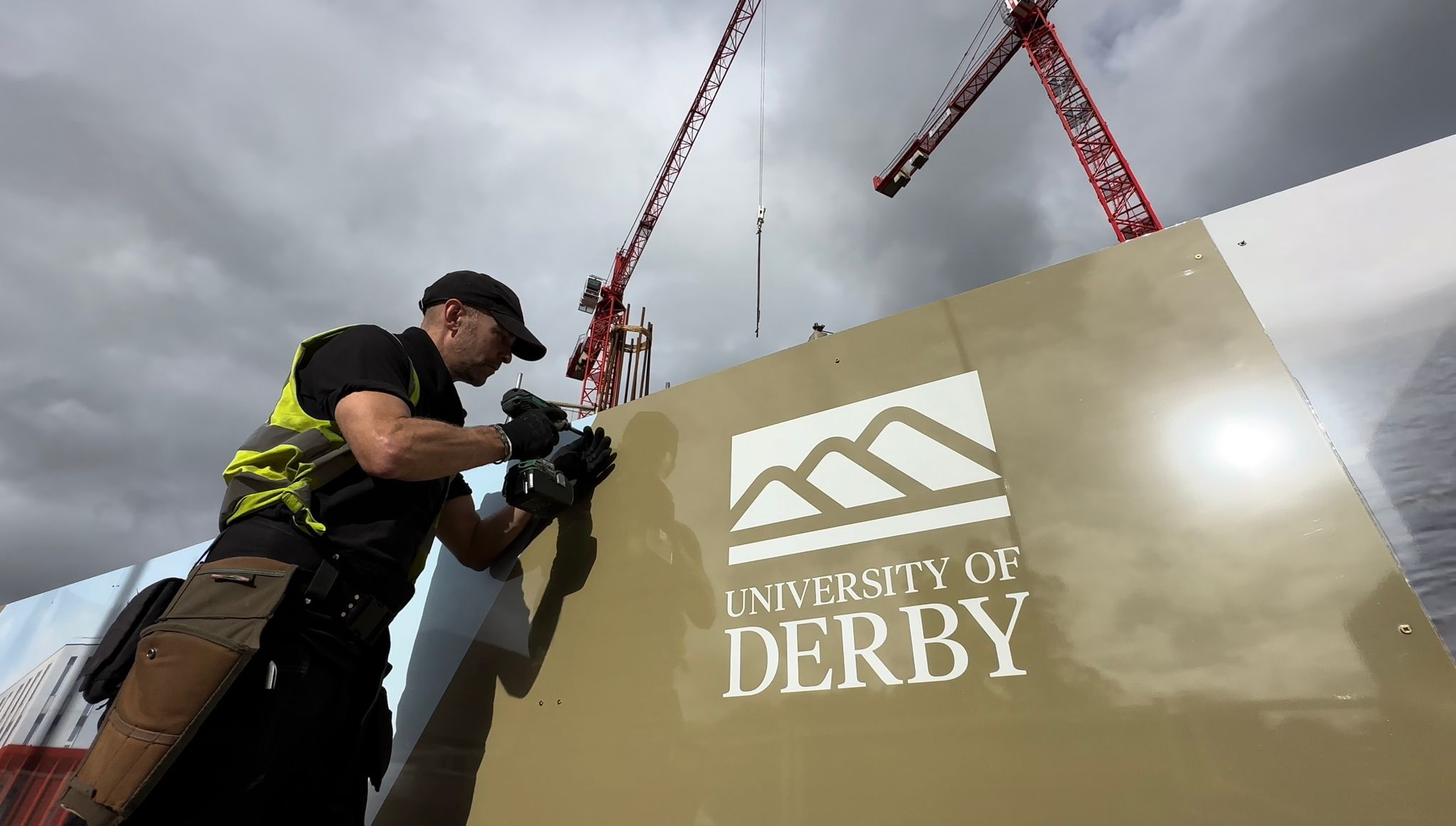 Construction worker in a high-visibility vest drills into a beige panel as red cranes loom overhead against a cloudy sky, with a University of Derby sign on the panel.