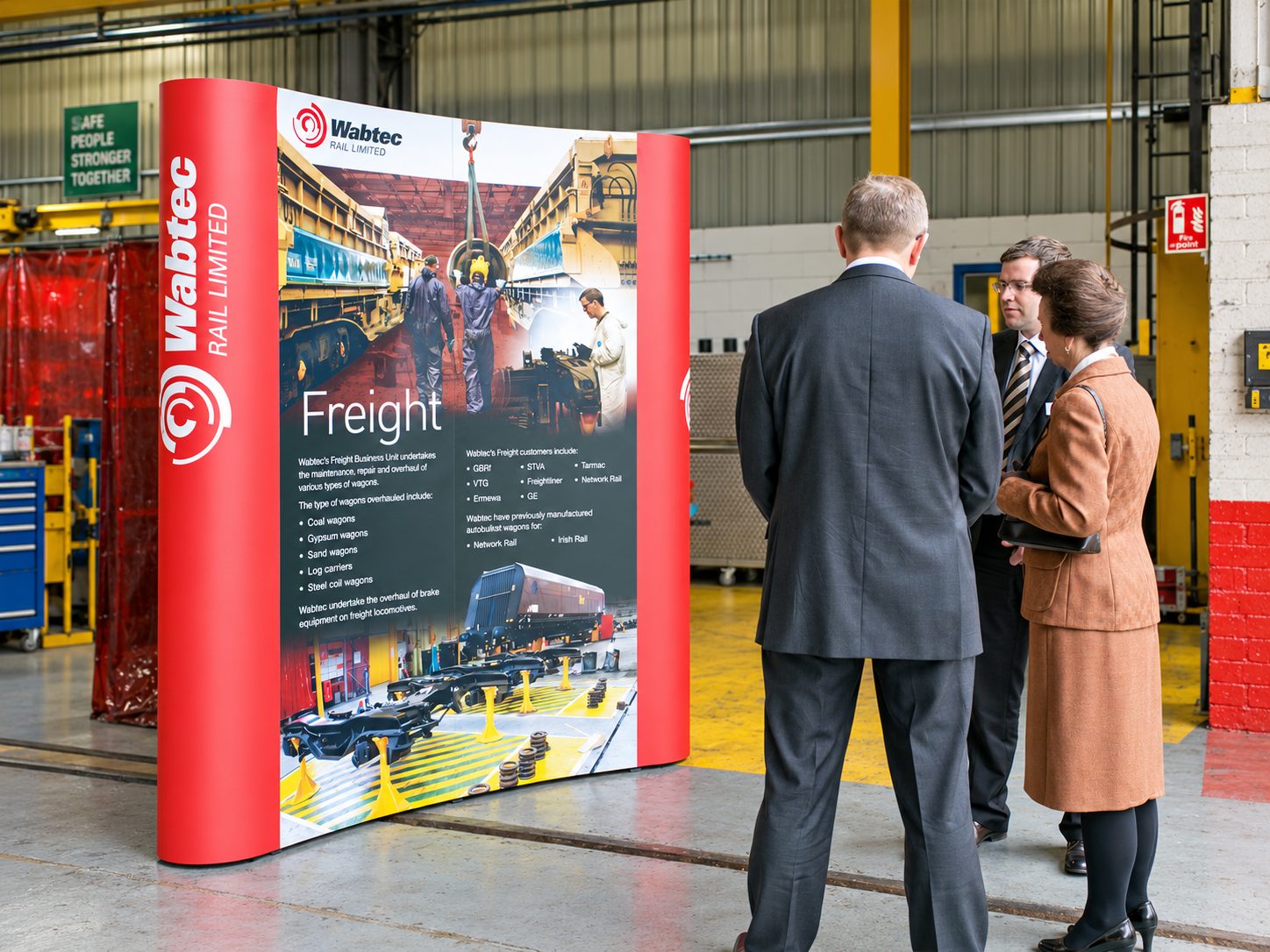 Promotional display for Wabtec Rail Limited about Freight services in a workshop, with three professionals reading the panel.