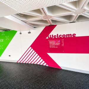 Colorful wall mural with green left panel saying 'terrace bar', bold pink diagonal stripe, and a 'Welcome' sign with a small directory plaque listing floors and venues.
