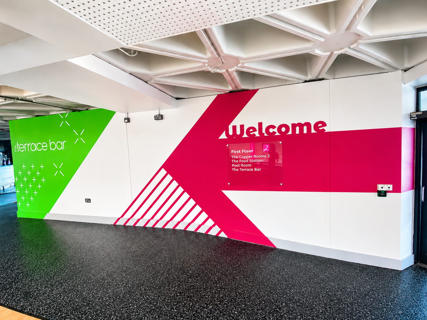 Colorful wall mural with green left panel saying 'terrace bar', bold pink diagonal stripe, and a 'Welcome' sign with a small directory plaque listing floors and venues.