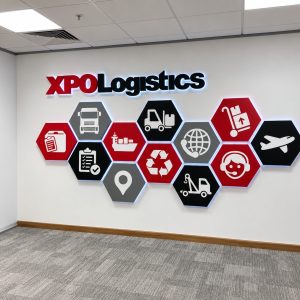 Sign reading 'XPO Logistics' on a white wall with red/black hex icons representing logistics services in a hallway.