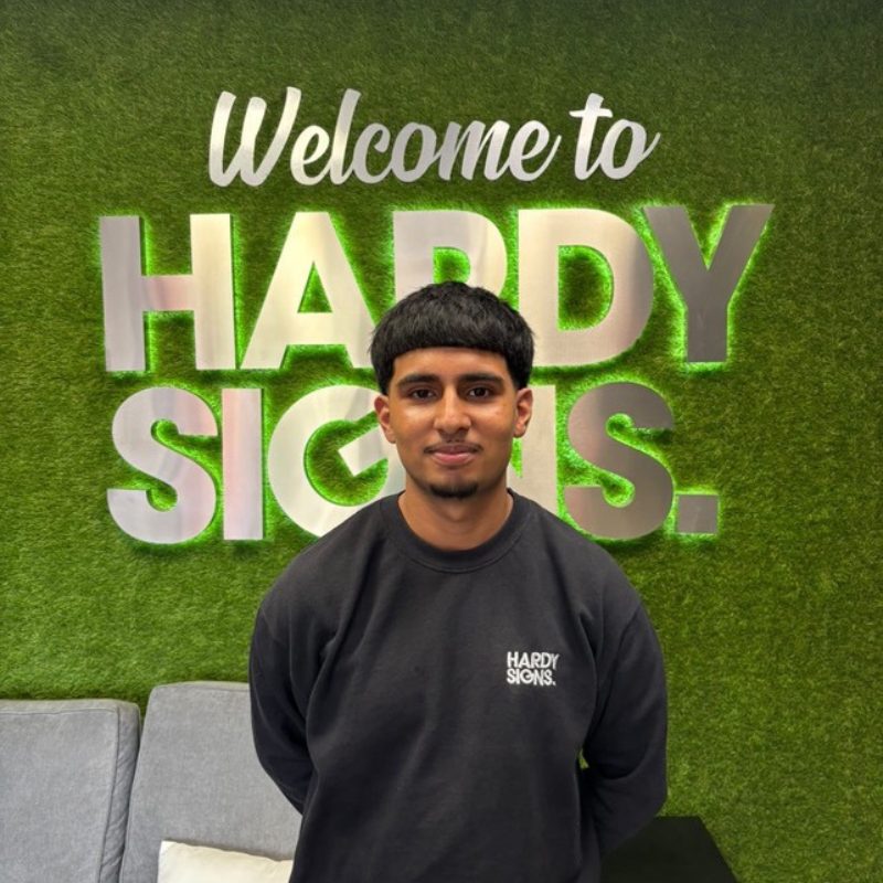 Young man in a black Hardy Signs sweatshirt stands in front of a green moss wall with large white letters reading 'Welcome to HARDY SIGNS'