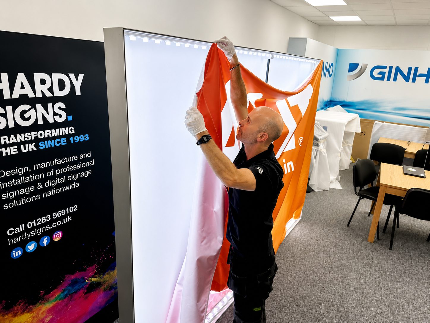 A man in a black polo with gloves unfolds an orange and white banner inside a signage showroom, with Hardy Signs branding on the left.