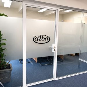 Office door with frosted glass panels and a black oval alba logo; a coat rack and potted plant on the left, blue carpeted floor in an office.
