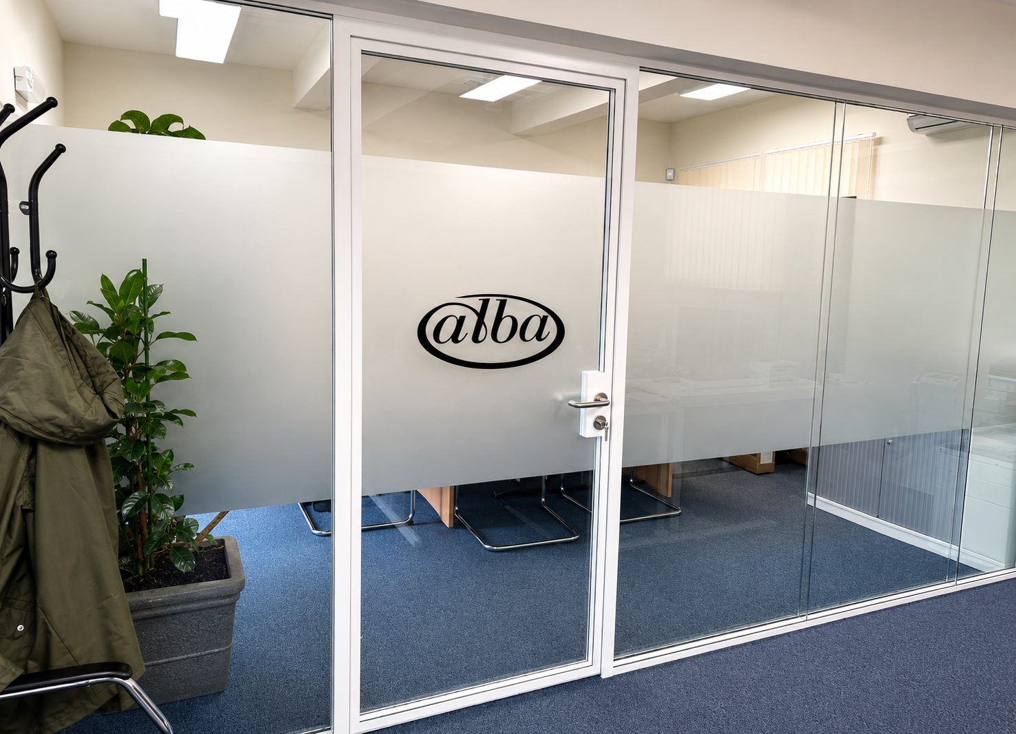 Office door with frosted glass panels and a black oval alba logo; a coat rack and potted plant on the left, blue carpeted floor in an office.