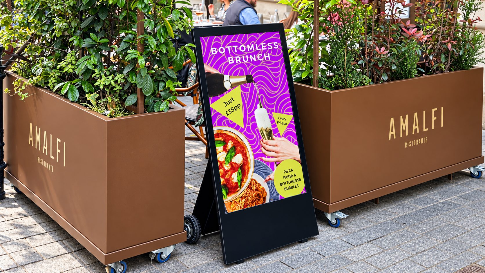 Poster advertising bottomless brunch with pizza and pasta, placed beside large brown planters at Amalfi Ristorante outdoor seating area.