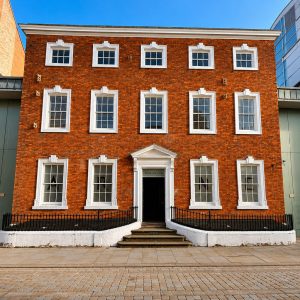 Three-story red brick building with white-framed sash windows and a central doorway, set between large green doors on a cobblestone plaza