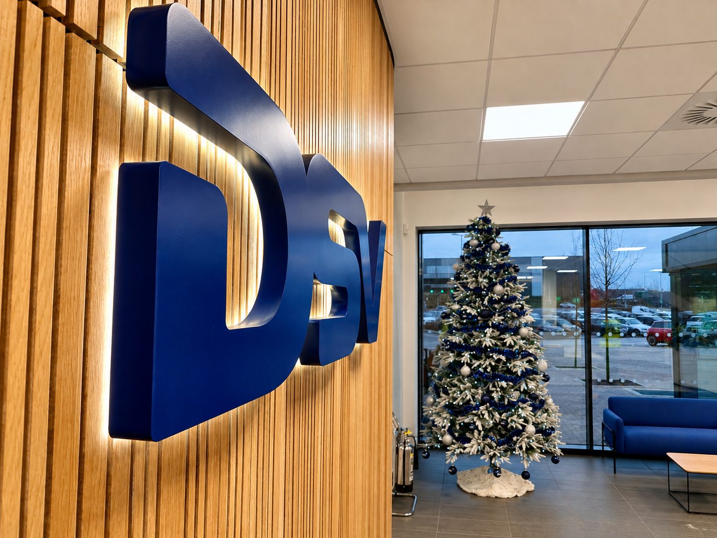 Office lobby with a large blue 3D logo on a wooden slat wall beside a decorated Christmas tree and seating visible through glass doors.