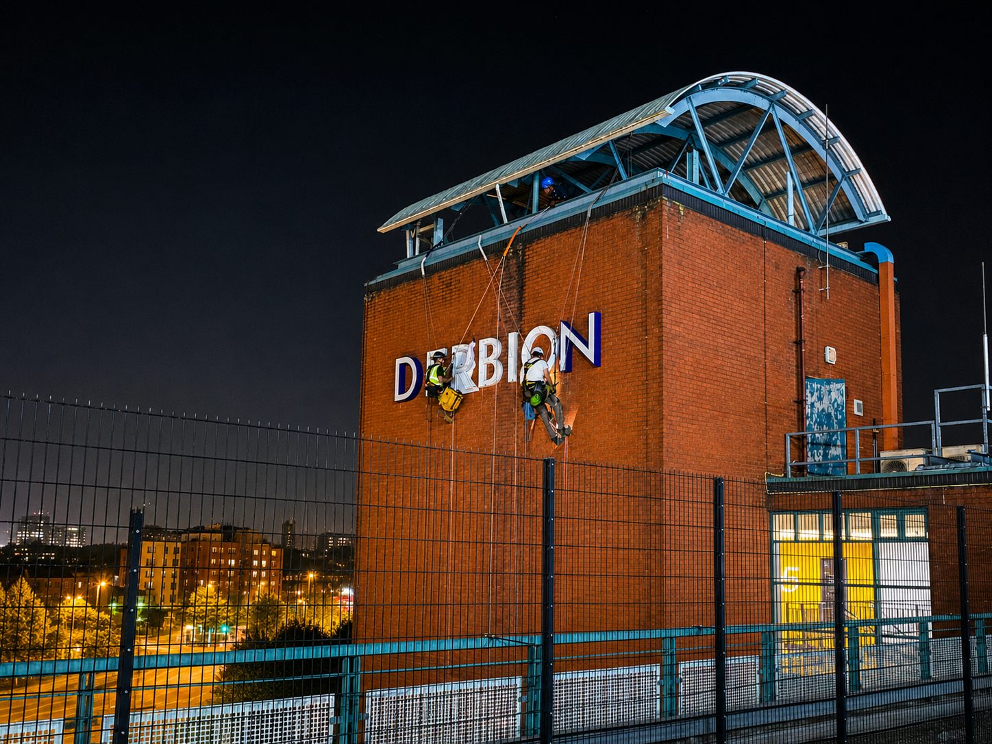 Two workers in safety gear rappelling down a brick building with large white letters on the wall at night.