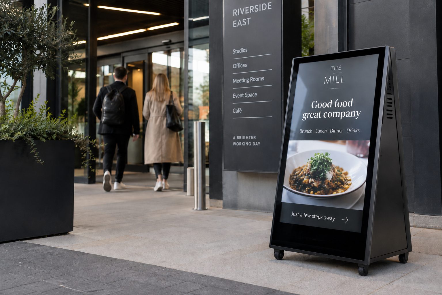 Outdoor street scene with a black electronic menu board advertising The Mill: Good food, great company; two people walk toward a building entrance.