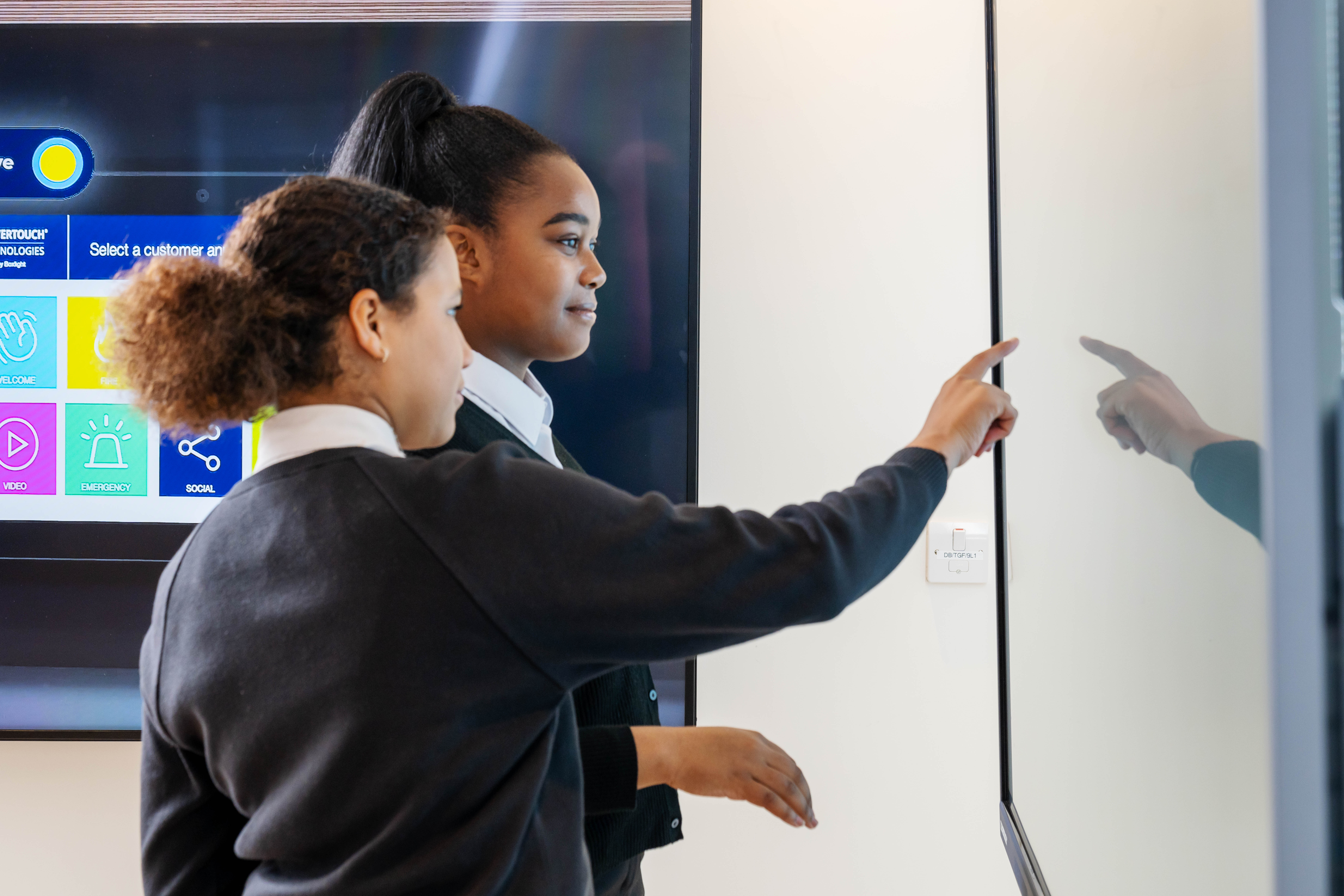 Two students in dark sweaters point at a large touchscreen displaying colorful app icons.