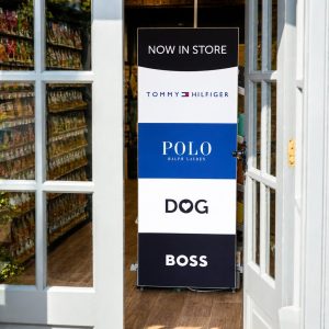 Store entrance with a tall vertical sign reading 'NOW IN STORE' and brand logos (Tommy Hilfiger, Polo Ralph Lauren, DOG, Boss) displayed in bold blocks.