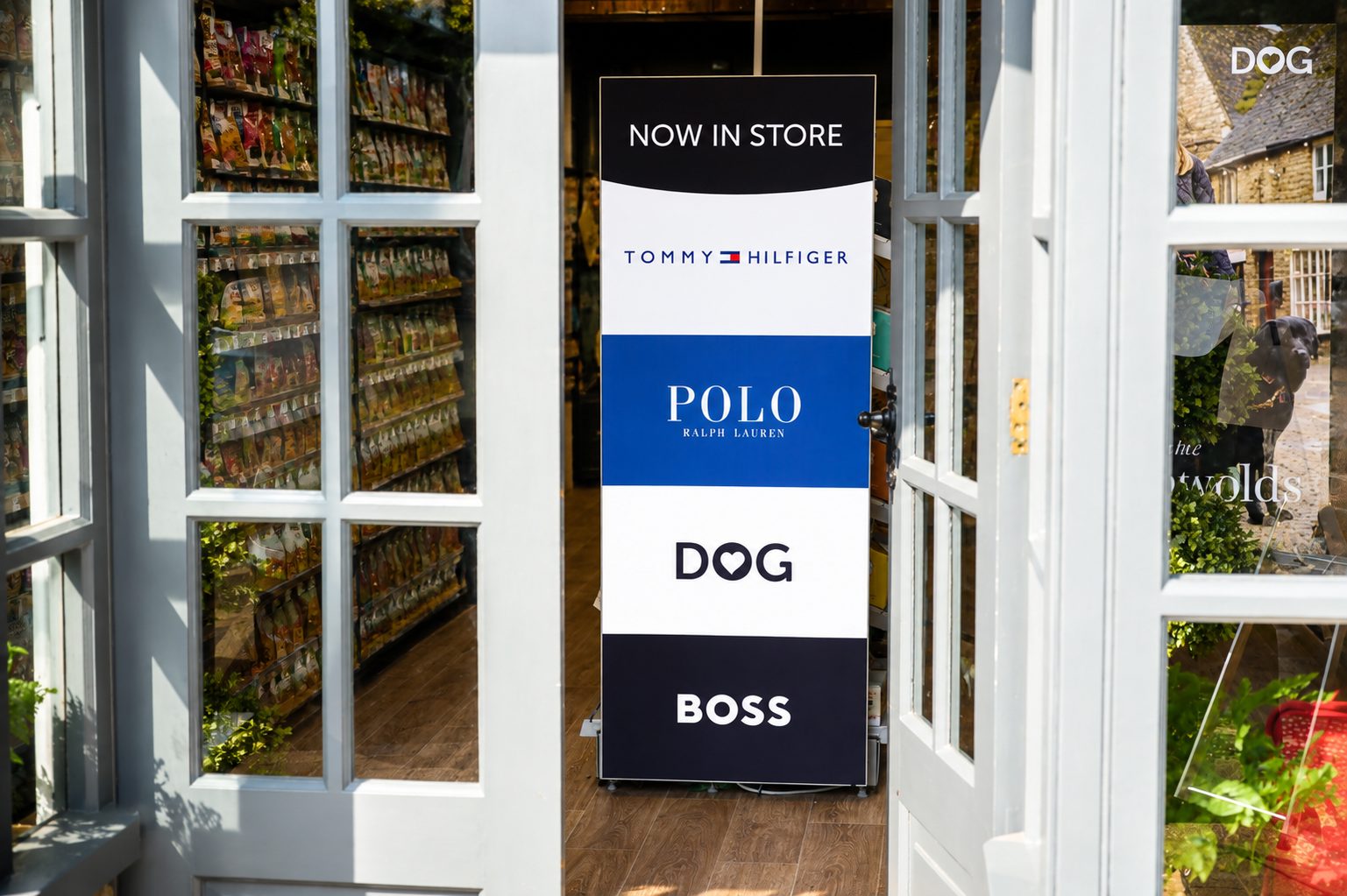 Store entrance with a tall vertical sign reading 'NOW IN STORE' and brand logos (Tommy Hilfiger, Polo Ralph Lauren, DOG, Boss) displayed in bold blocks.