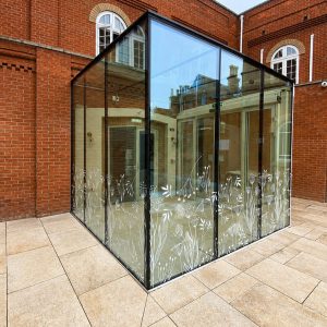 Glass-walled atrium extension with white floral etching along the bottom, set in a red-brick courtyard with beige tiled paving.